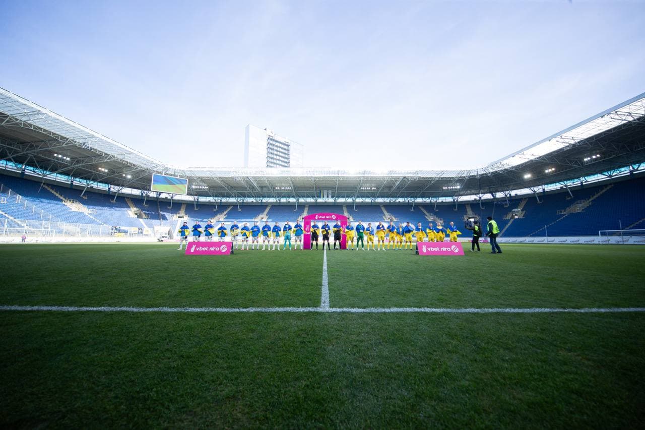 SC Dnipro-1 and FC Mynai lining up on the pitch at Dnipro Arena before kickoff, empty stands under martial-law spectator rules.
